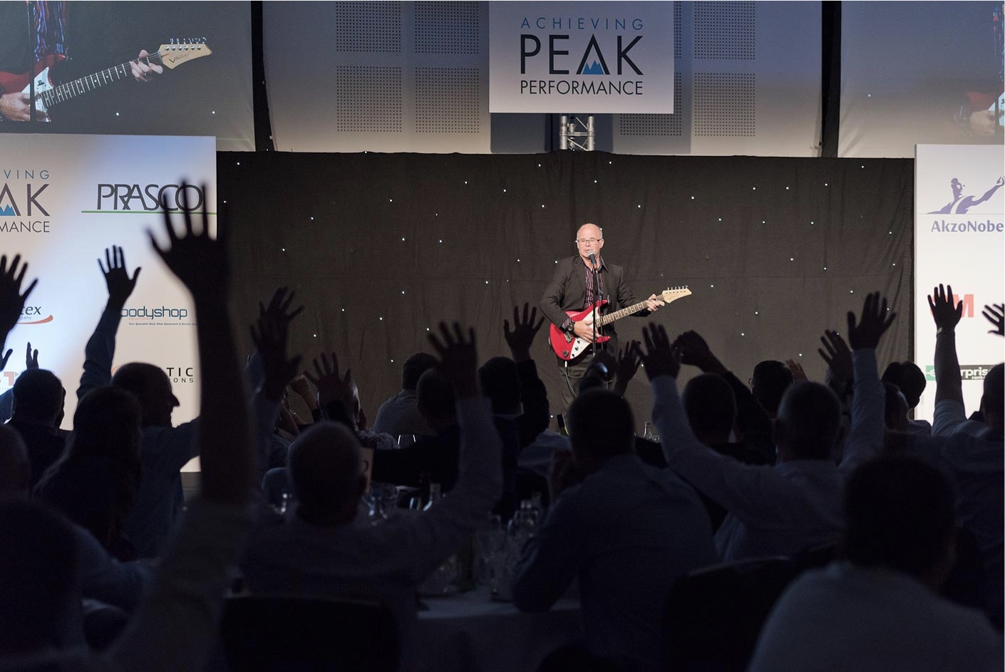 Andy Askins performing on the red guitar at a corporate event beneath an 'Achieving Peak Performance' banner, the audience with hands in the air